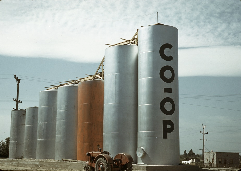 Large, cyndrilical grain elevators behind a tractor. The grain elevator in the foreground reads, 'Co-Op.'