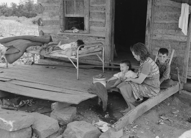 A young white woman holds and infant while one child sits on a broken porch while another lay on a cot. The porch appears unsafe and the cabin is very small. 