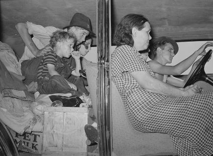A white family of five in a vehicle; the children in the back seat are balanced on boxes and other items. 