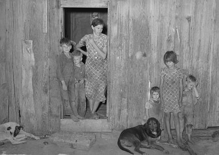 A white woman and two children in a doorway of a ramshackle house three more children and two dogs are in front of the house. 