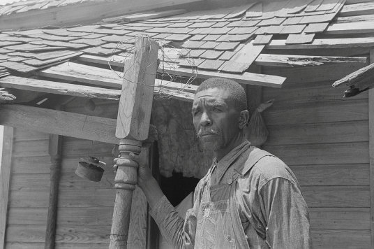 A Black man holds a post that is no longer attached to the roof over the porch. The roof is in significant disrepair. 