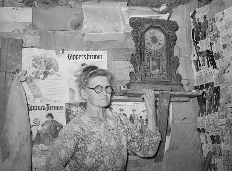 Older white woman standing in front of a wall insulated with magazine pages and cardboard. Her hand is touching a shelf holding an antique clock.  