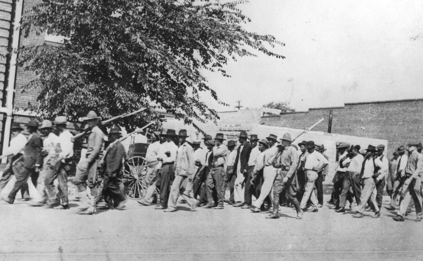 Black men walking down a street. A few armed white men are on the edge of the group.