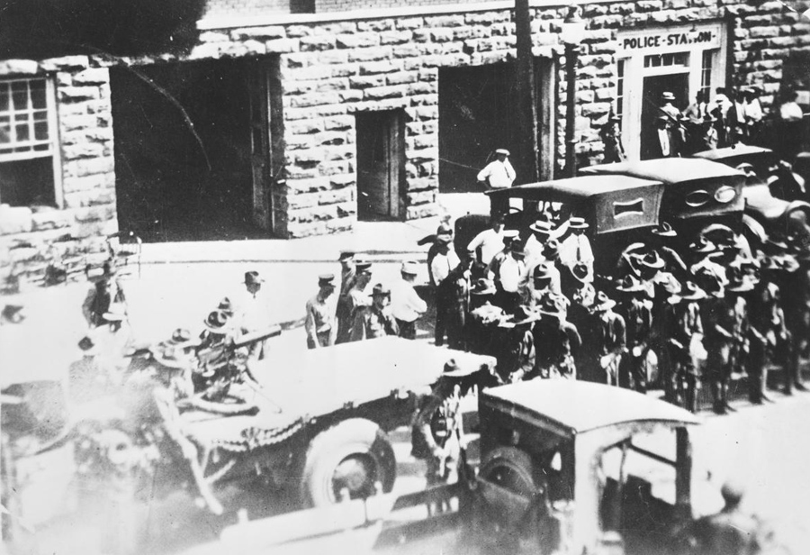 An overhead view of the street in front of a brick police station. Multiple uniformed men stand in the street.