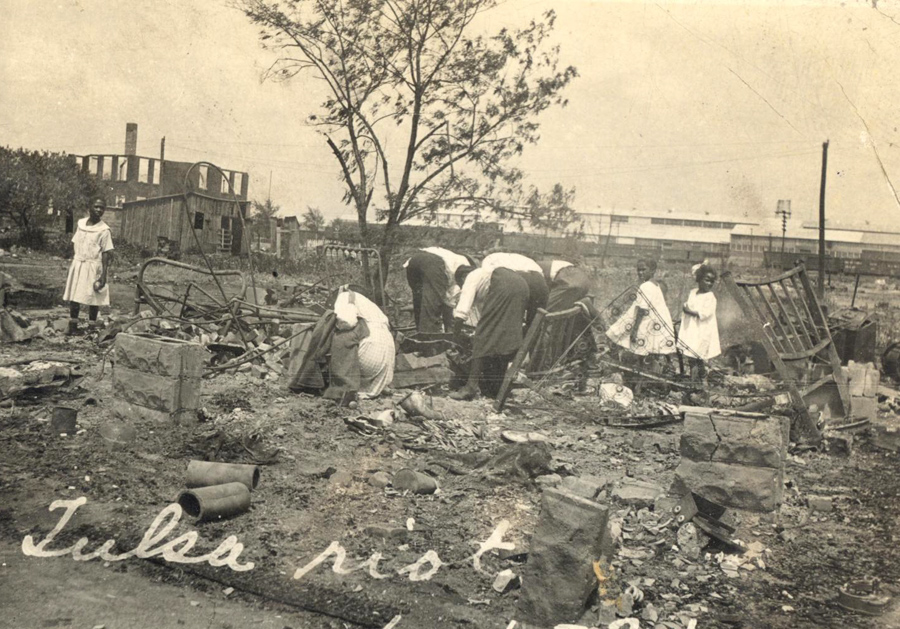 Black men, women, and children in the rubble. The men and women are bent down and looking through the remains. Three girls in white dresses stand by, and one looks at the camera.