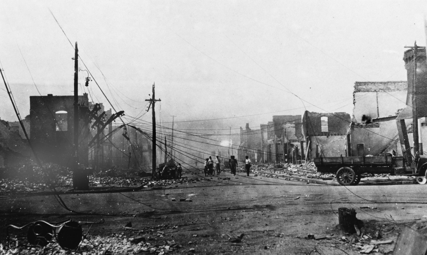 A small group walks down a street surrounded by burned out buildings and rubble. Electric wires and hardware hang down into the street. The remains of a vehicle are in the foreground.