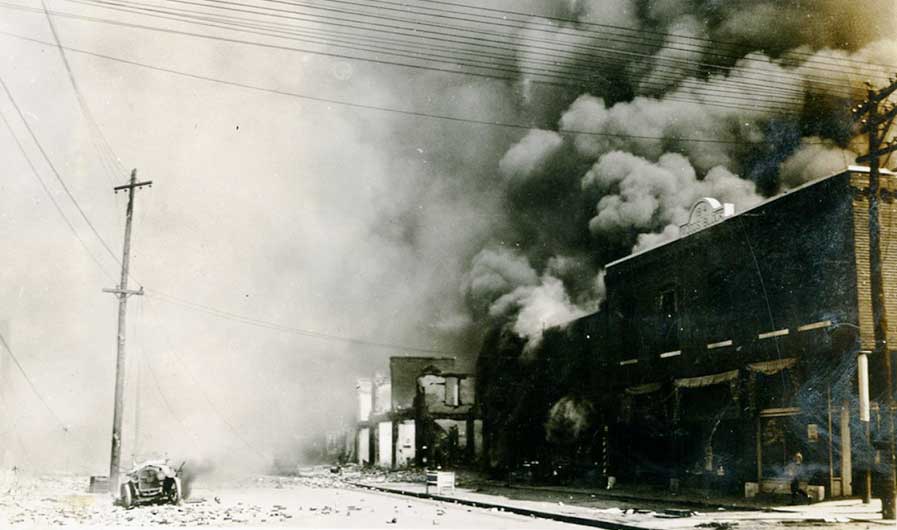 A burned out vehicle sits on an empty road. Across the street, smoke billows from a large brick building, which stands next to the rubble of other structures.
