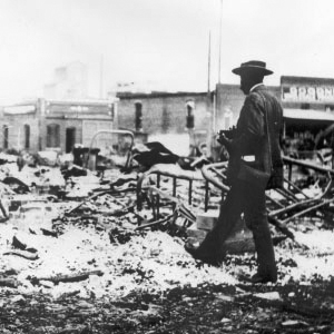 A Black man wearing a suit and holding a camera walks through the remains of a burned building
