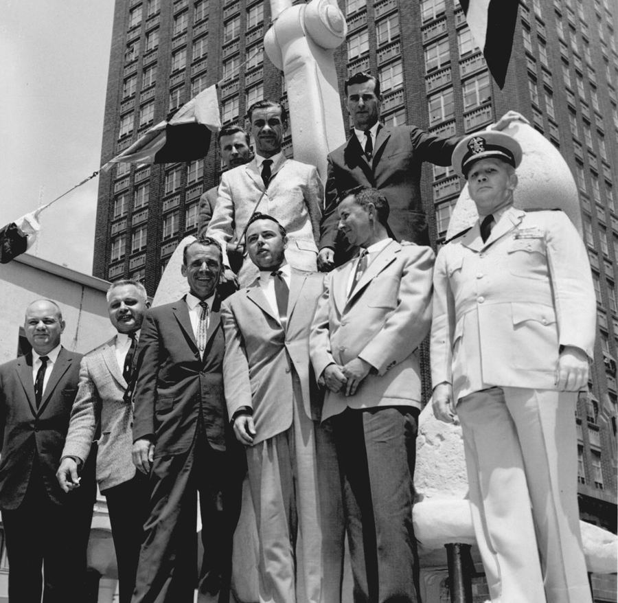 Men standing outdoors in front of an anchor recovered from the ship. A tall building is visible in the background.