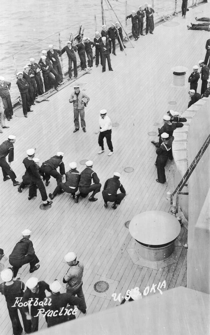 A row of sailors in a football formation on the a battleship's deck. Several men are lined up to watch the activity.