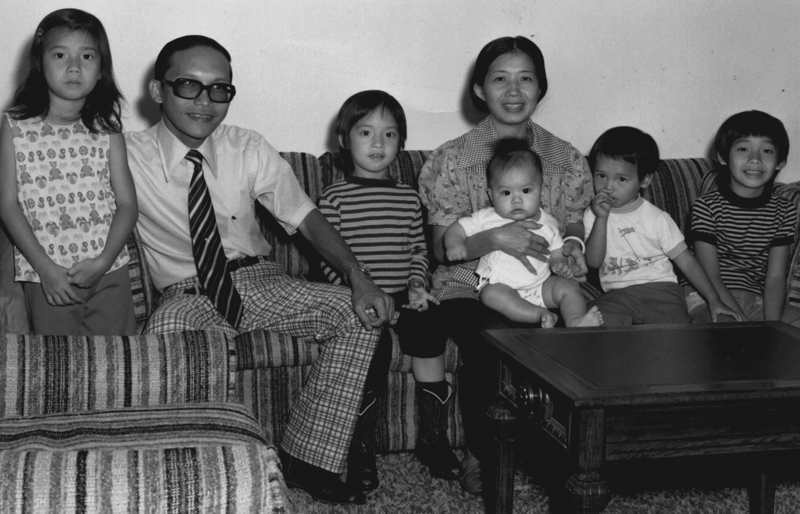 A Vietnamese father and mother holding a baby sit on a couch with their four young children. The parents smile at the camera. 
