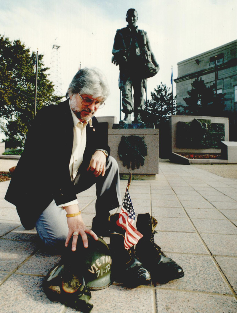 A man kneels on the ground resting his hands on a military helmet that reads 'Vietnam 71-72' and a pair of military boots. A small American flag stands inside the boot. A statue of an American soldier stands behind him. 