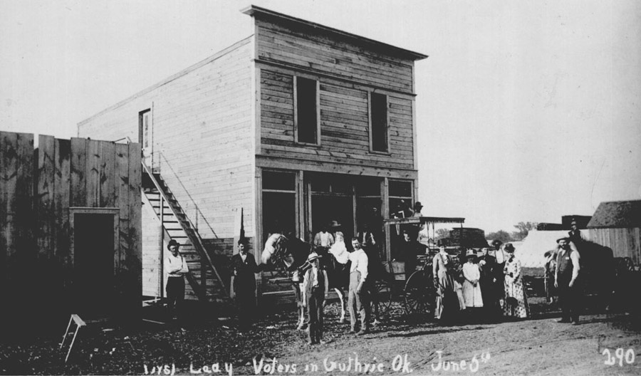 Four women, a few men, and a children in front of a two-story wood building
