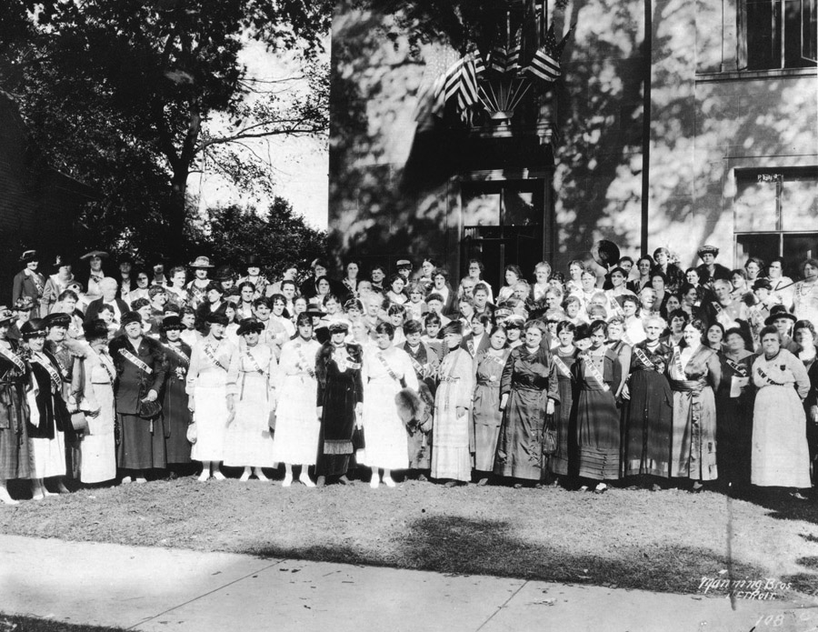 A large group of women stand outdoors for a photograph. Behind them is a large building with many American flags.