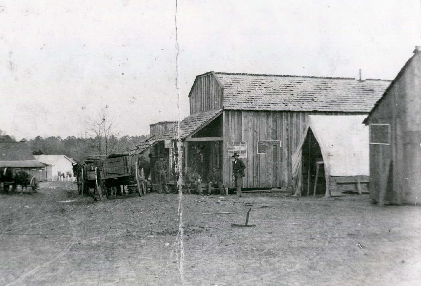 Historic view of Doaksville with wooden buildings, a tent, and a few horse-drawn wagons. A few people sit on the porch of one structure.