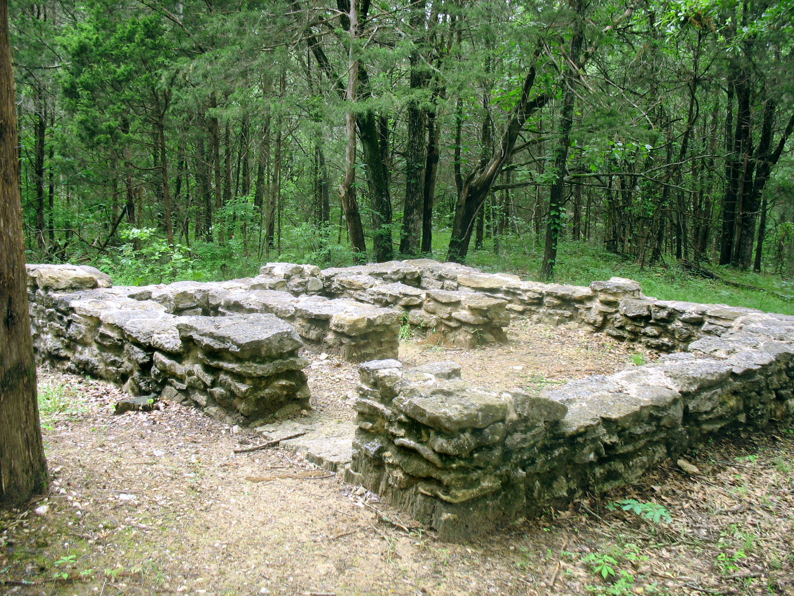 Short stone ruins of a building show where small rooms were located. A green heavily wooded area is in the background.