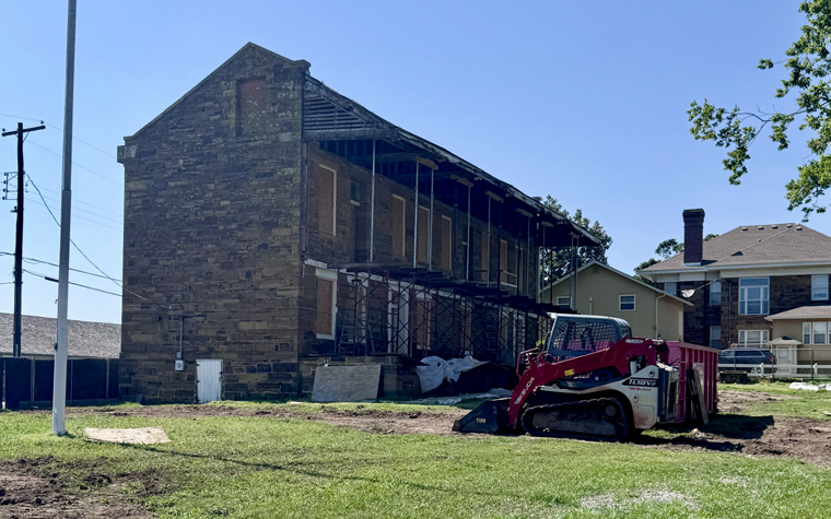 The two-story stone barracks under restoration, with boarded-up windows and a temporary balcony framework. A skid-steer loader sits nearby, next to a dumpster.