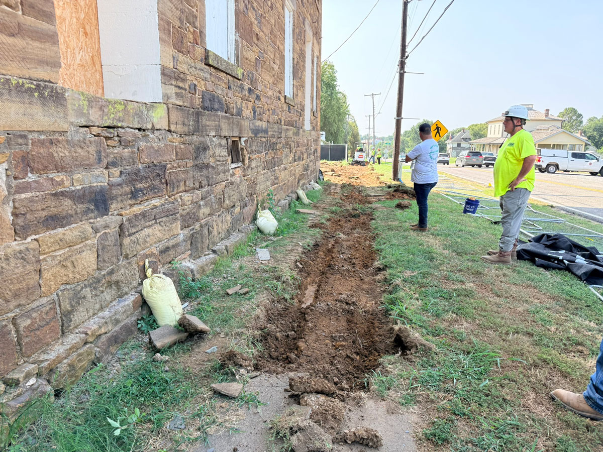 Two men stand outside the stone barracks, near a shallow trench. Cars and the historic hospital are in the background.