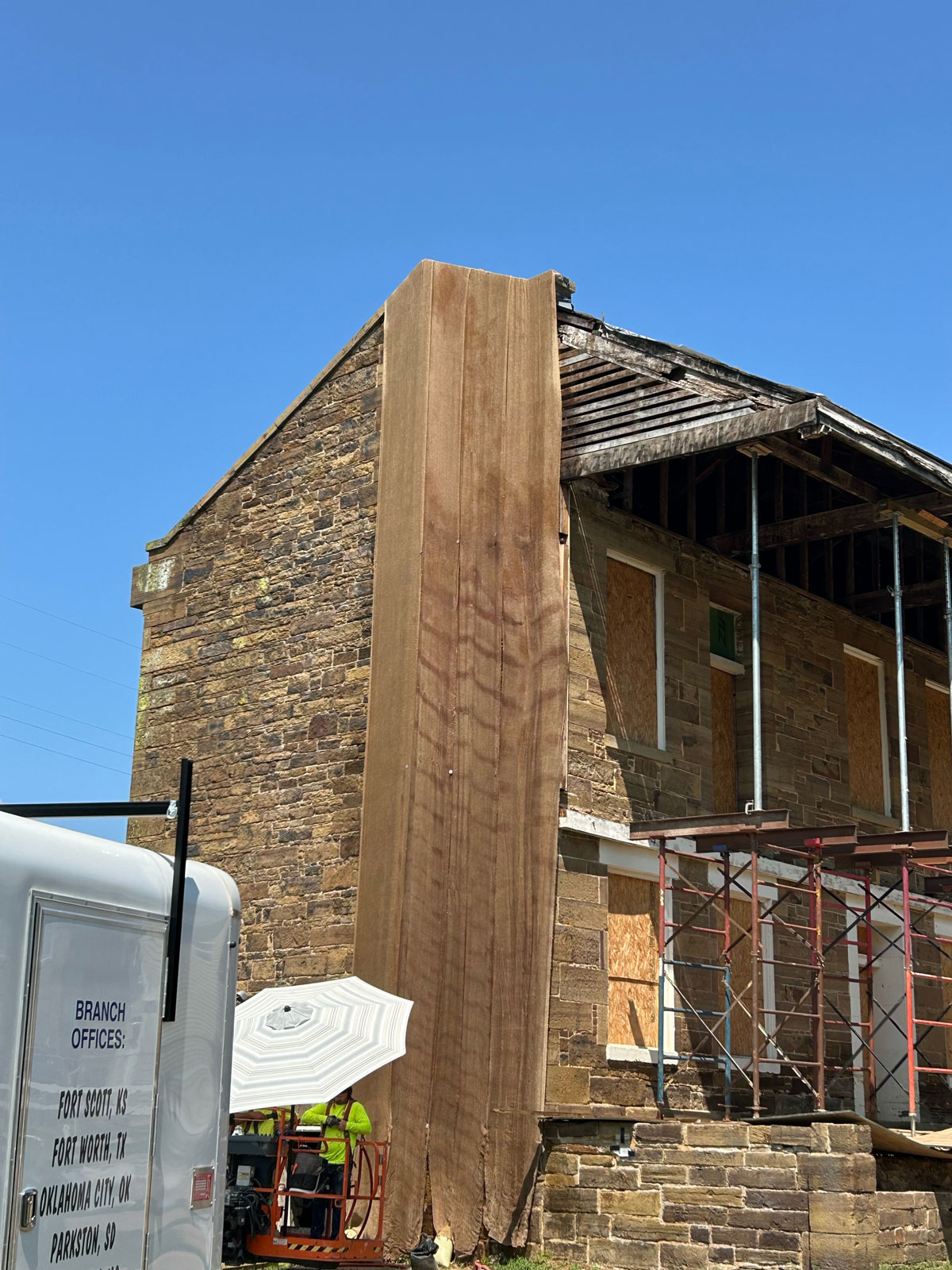A closer view of the burlap covering the end of the stone barracks. A small trailer and two workers under an umbrella are visible in the bottom-left of the photo.