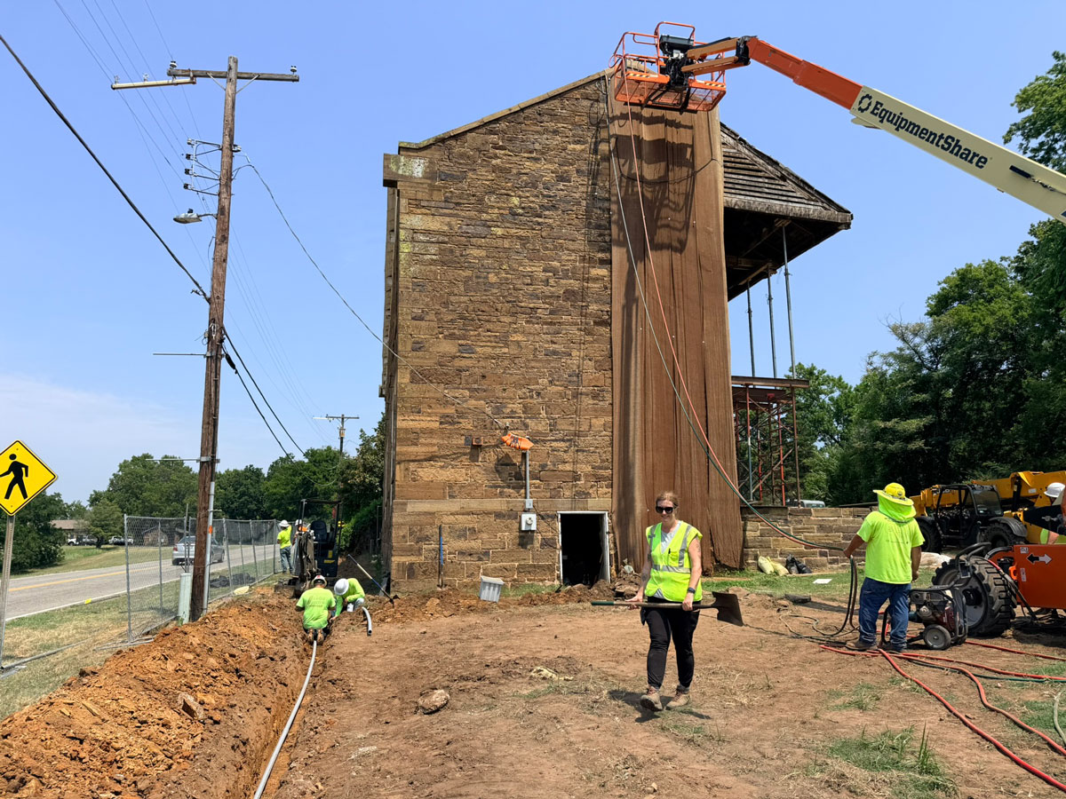 A view of the end of the barrackes shows a cherry picker at the top of the building. Long pieces of burlap cover the mortar from the top of the building to the ground. Workers in high-visibility clothing tend to a trench, one carries a shovel, and another looks toward the building.