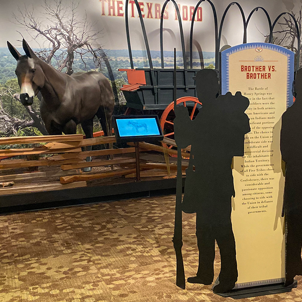 The sihlouette of a Civil War solider. He is holding a musket with a bayonet on the end and carrying supplies on his back. In the background is an exhibit featuring a mule and a wagon.
