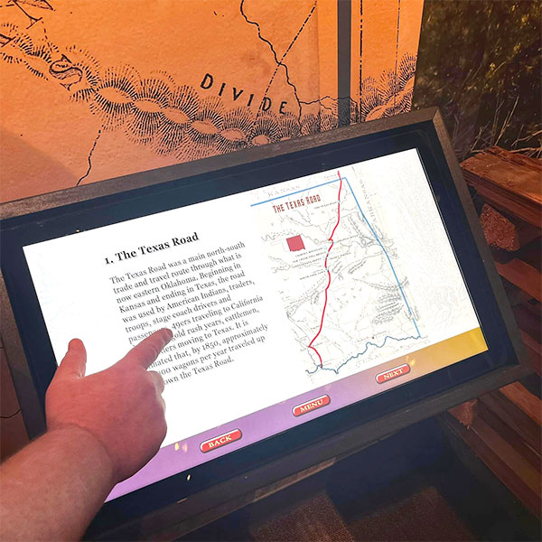 A hand points to an interactive touchscreen. On the screen is a small map and text about the Texas Road.
