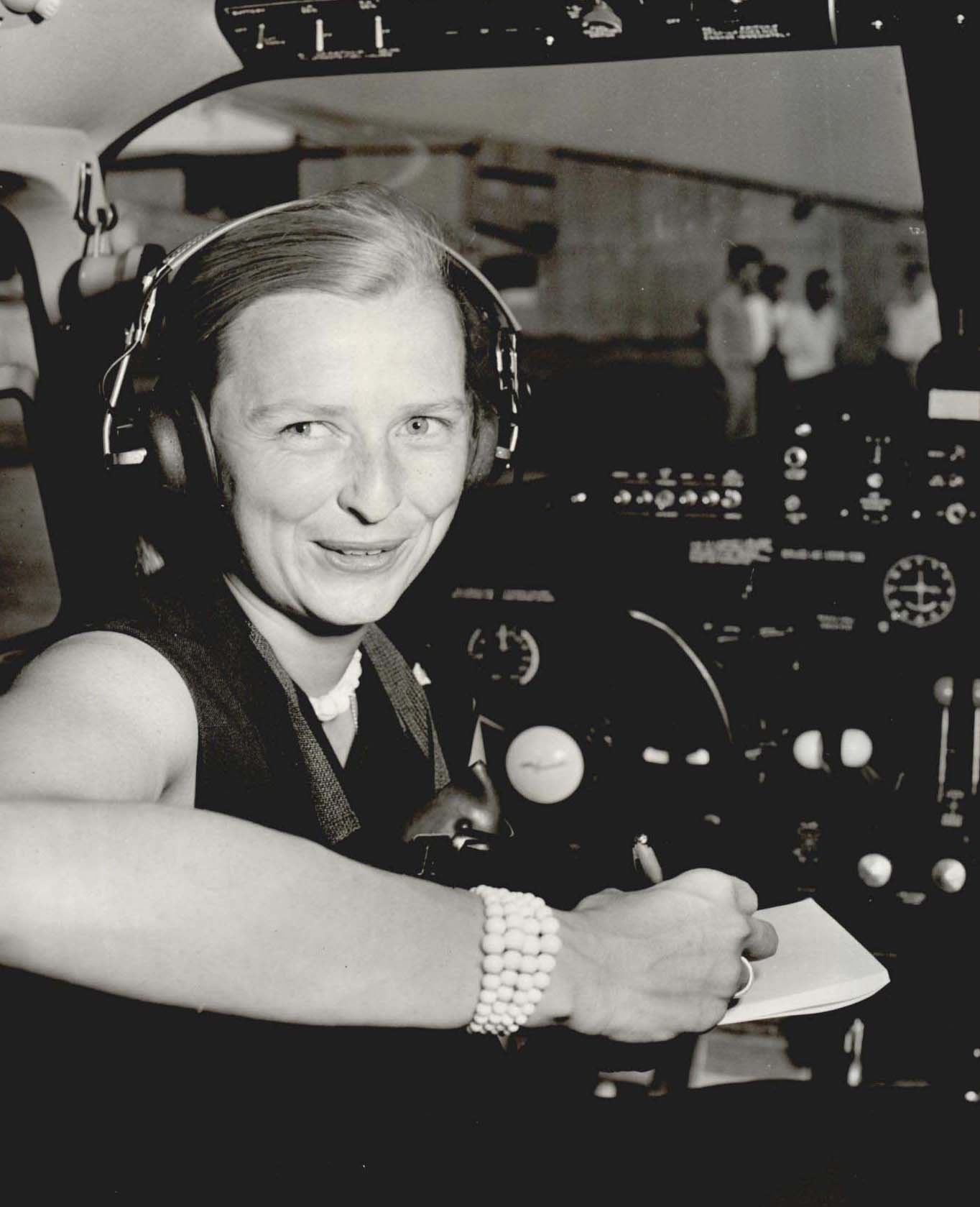 Aviator Jerrie Cobb sits in the cockpit of an aircraft wearing headphones as she smiles at the camera